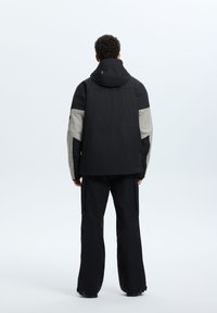 Giacca nera impermeabile con accenti grigi, cappuccio e una vestibilità comoda. Presenta una superficie liscia e dettagli rinforzati, abbinata a pantaloni neri.