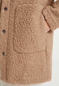 Abrigo marr�ón texturizado con una tela suave y peluda, gran bolsillo y botones a presión metálicos. El diseño es simple y funcional.