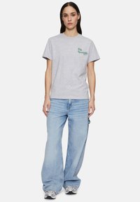 Camiseta de algodón gris con cuello redondo y mangas cortas, que presenta un logo verde de "Fila Sportslife". Combinada con unos pantalones vaqueros de pierna ancha de color azul claro.