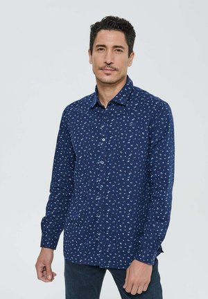 Homme portant une chemise à manches longues et à boutons de couleur bleu marine avec de petits motifs floraux blancs, se tenant devant un fond clair uniforme.
