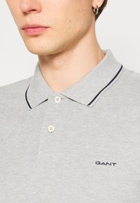 Pilkas polo marškinėlis, pagamintas iš tekstūruotos medžiagos, su dviem mygtukų užsegimu, apykakle su mėlyna juostele ir mažu siuvinėtu logotipu.