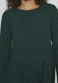 Abito a maniche lunghe con scollatura tonda, realizzato in tessuto verde scuro testurizzato, con vita arricciata e sottili righe orizzontali.