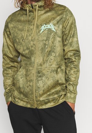 Man draagt een groene gemêleerde hooded trui met ritssluiting en trekkoorden, met een gestileerd logo op de borst, handen in de zakken, zwarte broek zichtbaar eronder.