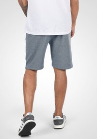 Egy férfi kék sortot, fehér pólót és szürke Adidas sportcipőt viselve háttal áll a kamerának egy fehér háttér előtt.