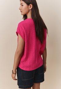 Blouse rose vif texturé à manches courtes, associée à un short en denim foncé. Coutures apparentes sur le short, modèle vu de dos.