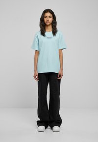Ljusblå oversized t-shirt gjord av mjukt tyg, med en liten svart logotyp. Kombineras med svarta vida byxor och vita skor.