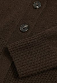 Brun stickad cardigan med ribbade ärmslut, prydd med två mörka, texturerade knappar. Tyget ser mjukt ut med en jämn väv.