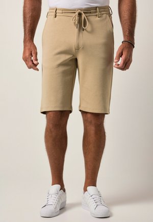 Mand iført beige snoretræks-shorts og hvide sneakers stående mod en ensfarvet lys baggrund.
