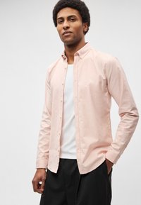 Ροζ ανοιχτό ροζ πουκάμισο από λείο ύφασμα, με κλασικό γιακά, μακριά μανίκια και ίσια γραμμή. Φοριέται πάνω από λευκή μπλούζα.