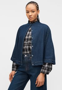 Γυναίκα με πλεξούδες που φοράει σκουρό μπλε τζιν κάπα-τζάκετ πάνω από μαύρο και λευκό καρό πουκάμισο και ψηλόμεσα σκουρό μπλε τζιν παντελόνια.