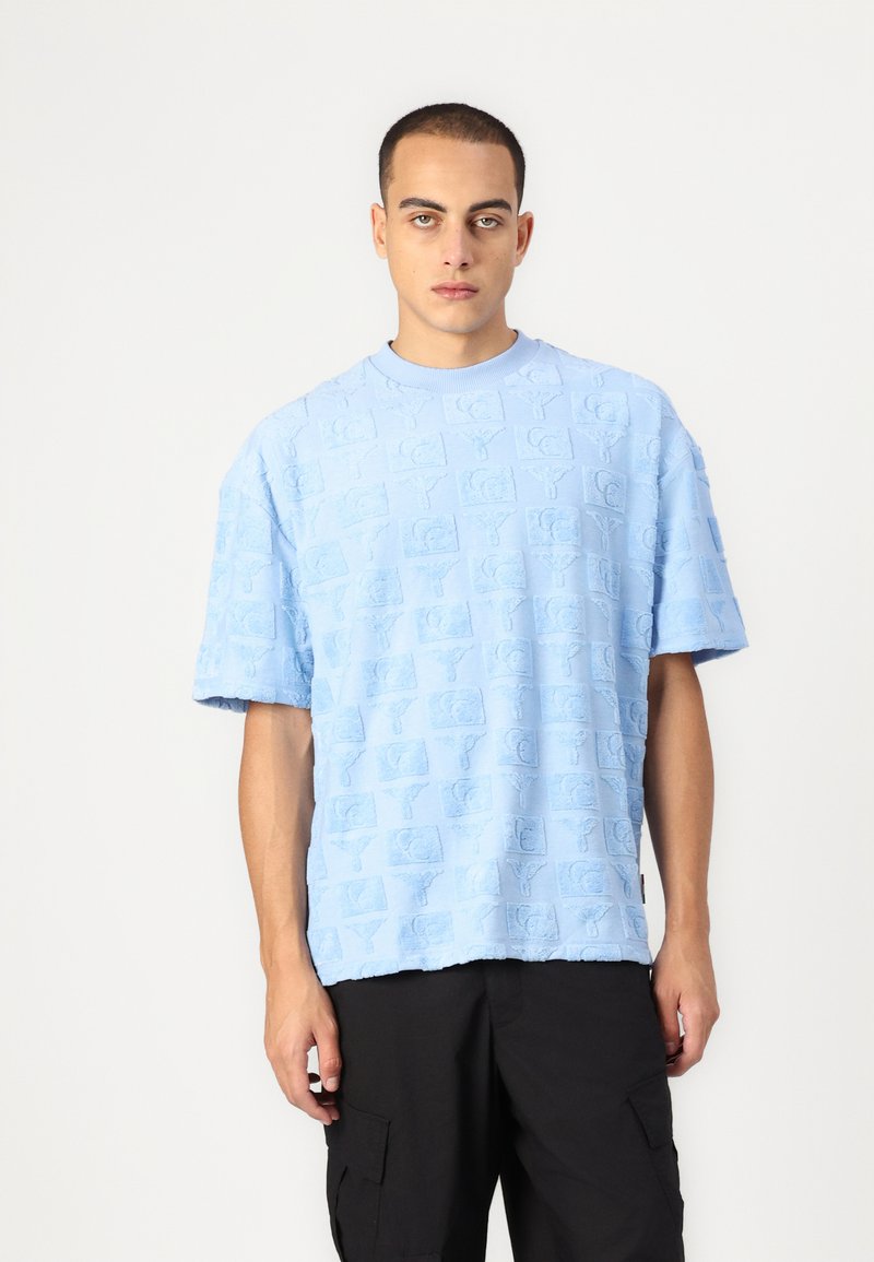 Lyseblå t-skjorte med kort erm og teksturert, hevet logomønster. Rund hals, avslappet passform, kanten sitter litt under livet. Svarte cargo-shorts.