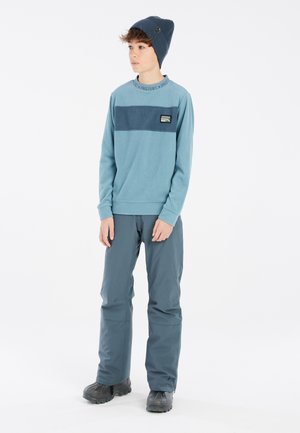 Sweat-shirt en polaire bleu clair avec une bande horizontale bleue marine, écusson logo ; associé à un pantalon bleu foncé et des bottes noires ; bonnet simple.