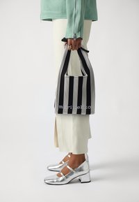 Striped black and white tote bag with a soft texture, held by a person wearing a mint green jacket and cream-colored pants, with silver shoes.