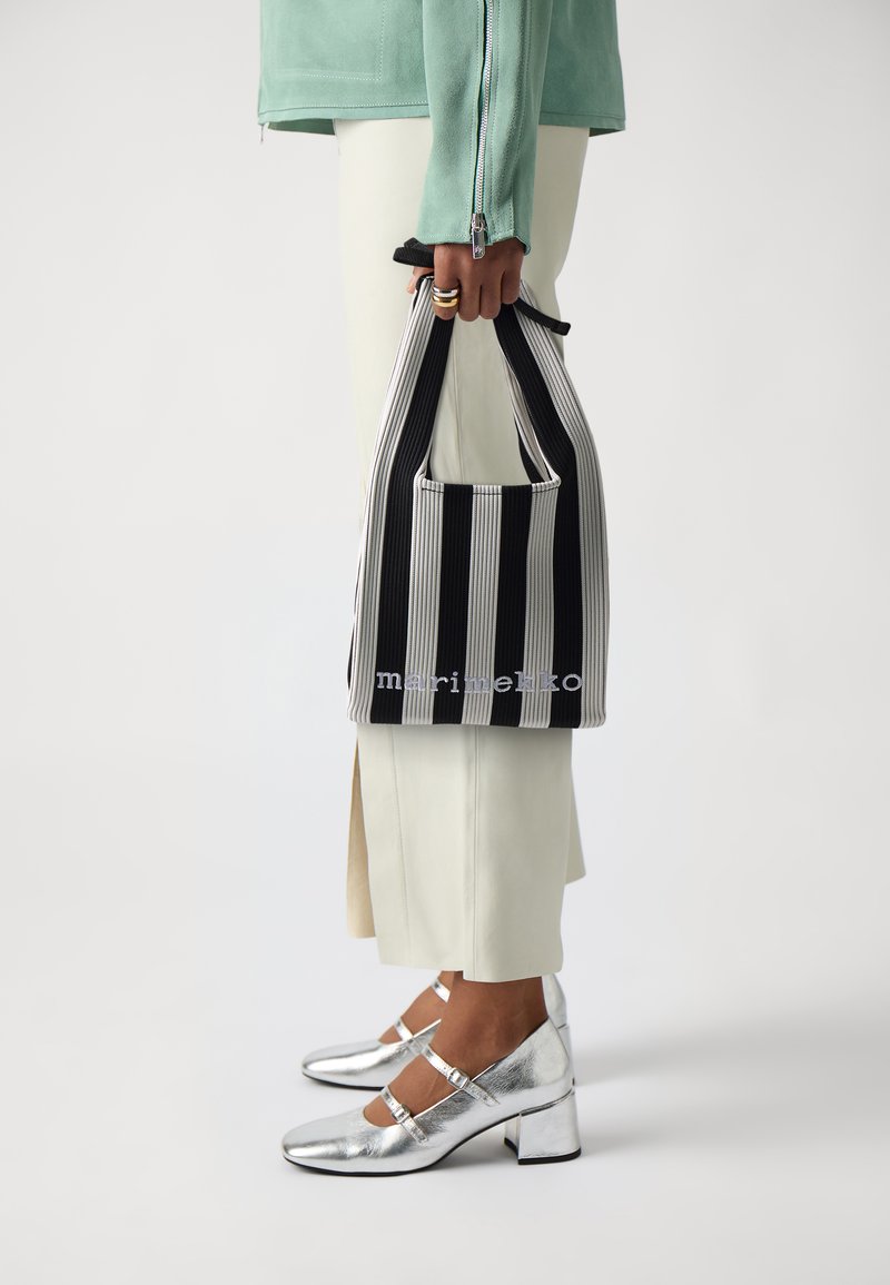 Striped black and white tote bag with a soft texture, held by a person wearing a mint green jacket and cream-colored pants, with silver shoes.