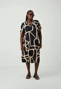 Žena u haljini do koljena crne boje s bež apstraktnim geometrijskim uzorcima, crnim sandalama s visokom petom, sunčanim naočalama i srebrnim privjeskom, stoji.