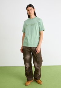 Ανοιχτό πράσινο βαμβακερό t-shirt με ανάγλυφο λογότυπο "Timberland", συνδυασμένο με καφέ cargo παντελόνι που διαθέτει πολλές τσέπες και άνετη εφαρμογή.