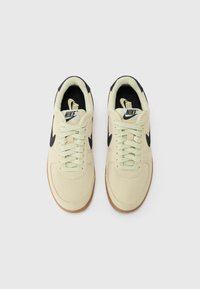 Nike sneakers har en lys cream canvas overdel, sorte accenter, gummisål og cream farvede snørebånd, designet med en glat tekstur og strømlinet form.
