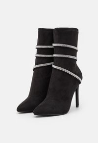 Bottes à talon aiguille noires en daim, à bout pointu, avec trois bandes en strass argentés enroulées autour de la tige, sur fond blanc.