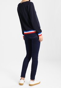 Navyblaues Langarmoberteil mit rundem Ausschnitt und gestreiftem Saum in Rot, Weiß und Blau, kombiniert mit dunklen Skinny-Jeans und weißen Turnschuhen.