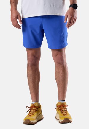 Uomo che indossa pantaloncini atletici blu, calzini bianchi, scarpe da trail running gialle e un orologio da polso nero, in piedi contro uno sfondo semplice.