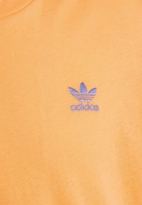 Oranžové bavlněné tričko se štíhlým výšivkovým logem Adidas v purpurové barvě se třemi pruhy připomínajícími listy. Měkká textura s designem kulatého výstřihu.