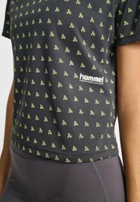 Svart sporttröja med gröna mönstrade detaljer. Har andningsbart tyg och en figurnära design. Logotyp visas längst ner till vänster.