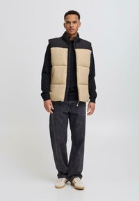 Vaatteena on beige ja musta pehmustettu liivi, jossa on korkea kaulus ja vetoketjusuljenta. Se on päällä mustan neuleen, tummien farkkujen ja valkoisten lenkkareiden kanssa.