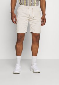 Uomo che indossa pantaloni corti beige con cordino, calze bianche alla caviglia, sneakers bianche e una camicia con fantasia, in piedi su uno sfondo neutro.