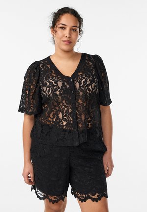 Frau trägt schwarze, florale Spitzenbluse mit Knöpfen, Puffärmeln und passende Spitzen-Shorts, steht vor weißem Hintergrund.