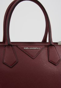 Bordeauxfarvet tote-taske med struktureret syntetisk materiale, dobbelte håndtag og sølvlogo. Har trekantede accenter på siderne.