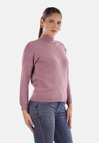 Donna con capelli scuri raccolti in un ponytail, che indossa orecchini d'oro, un maglione a collo alto rosa e jeans blu, rivolta di fronte con un'espressione neutra.