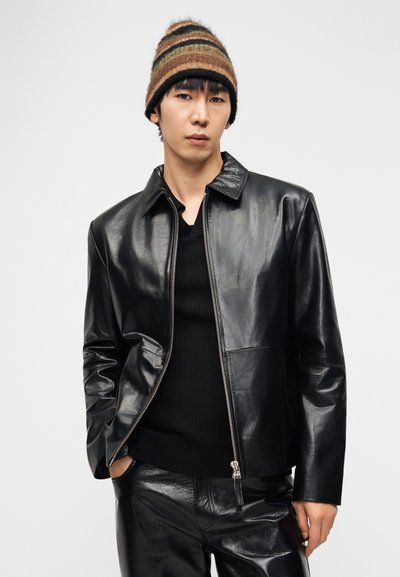 Νέος άνδρας ντυμένος με μαύρο δερμάτινο μπουφάν και παντελόνι, φορώντας ριγέ πλεκτό σκουφί καφέ, πράσινου και μαύρου, στέκεται με τα χέρια εν μέρει στις τσέπες.