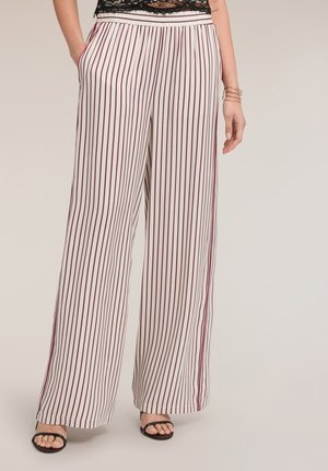 Frau trägt weiße, weite Hosen mit dünnen, vertikalen roten Streifen, schwarze Riemchensandalen und ein schwarzes Spitzenoberteil, steht mit einer Hand in der Tasche.