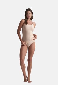 Nude badpak in glad weefsel, met een vierkante halslijn, dunne banden en een gematigde uitsnede. Model past de zijkant aan terwijl ze staat.