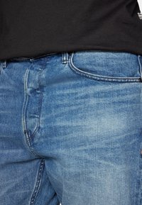 Kék farmernadrág közepes árnyalatban, enyhén kopott és koptatott felülettel. Klasszikus öt zsebes kialakítással és cipzáras záródással rendelkezik.