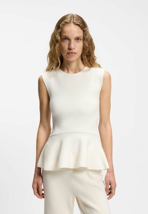 Frau mit welligem blondem Haar, die ein ärmelloses weißes Peplum-Top und passende weiße Hosen trägt, vor einem schlichten hellen Hintergrund stehend.