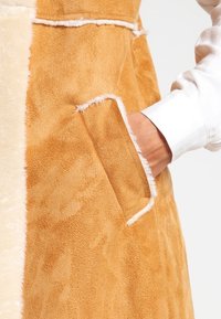 Tan faux fur-väst med en mjuk textur, en krämfärgad panel och en sidoficka med kontrasterande vit trim.