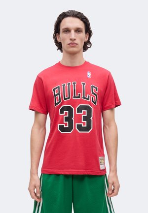 Mladý muž s tmavými kučeravými vlasmi, ktorý nosí červené tričko Chicago Bulls s číslom 33 a zelené basketbalové šortky, stojí na svetlom pozadí.