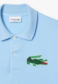 Ljusblå pikétröja med texturerad tyg, som har en grön broderad krokodil-logotyp på vänster sida. Klassisk passform med knappstängning.