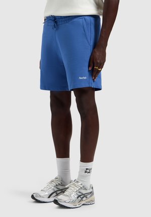 Persoon draagt blauwe PurePath-short, witte merkensokken en zilveren hardloopschoenen, staand tegen een effen achtergrond.