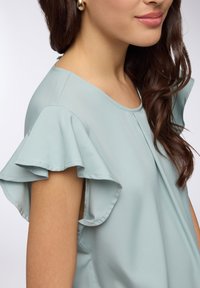 Blusa azzurra chiara con maniche a volant, scollatura rotonda e tessuto morbido e liscio. Design minimalista senza motivi o dettagli visibili.