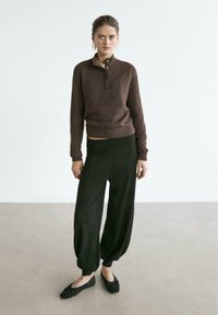 Brauner, gestrickter Pullover mit hohem Kragen und Knopf-Detail, kombiniert mit lockeren schwarzen Hosen und schwarzen flachen Schuhen. Glatte Textur, entspannte Passform.