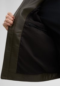 Innenfutter einer olivgrünen Lederjacke mit schwarzer Besatz, ausgestattet mit einer Reißverschlusstasche für sicheren Stauraum und sichtbaren Nahtdetails.
