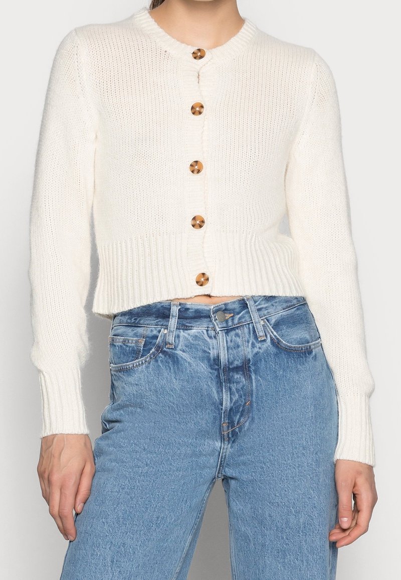 Cardigan en tricot crème avec un col rond, doté de six boutons en bois, de poignets côtelés et d'une silhouette raccourcie, associé à un jean bleu clair.