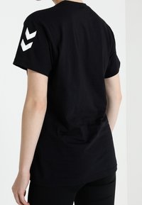 Camiseta de algodón negra de manga corta, con un cuello redondo y dos patrones en chevron blancos en el hombro izquierdo. Espalda lisa.