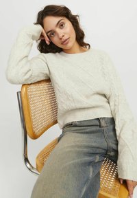 Maglione beige chiaro a coste con orlo a coste, indossato con jeans di denim a vita alta. Seduto su una sedia di legno con schienale intrecciato.