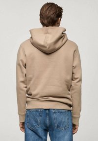 Persona di spalle che indossa un hoodie beige e jeans blu, in piedi contro uno sfondo grigio chiaro uniforme.