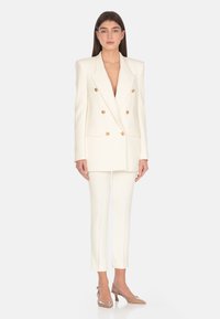 Blazer bianco doppiopetto con bottoni dorati, abbinato a pantaloni bianchi aderenti e décolleté beige. Design sartoriale, texture elegante e linee pulite.