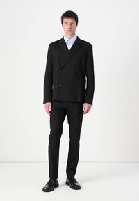 Schwarze doppelt knöpfbare Jacke mit einem strukturierten Muster, versehen mit großen runden Knöpfen und einem Reverskragen, kombiniert mit schwarzen Hosen und Schuhen.