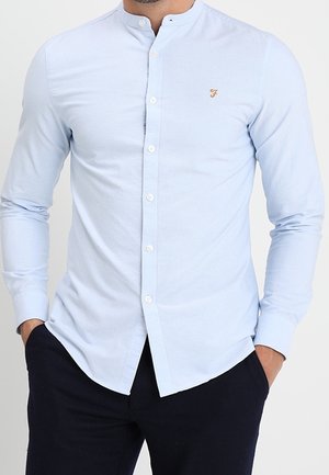 Mann trägt ein hellblaues Hemd mit Mandarinkragen und marineblaue Hose, Hände in den Taschen, steht vor einem einfarbigen Hintergrund.